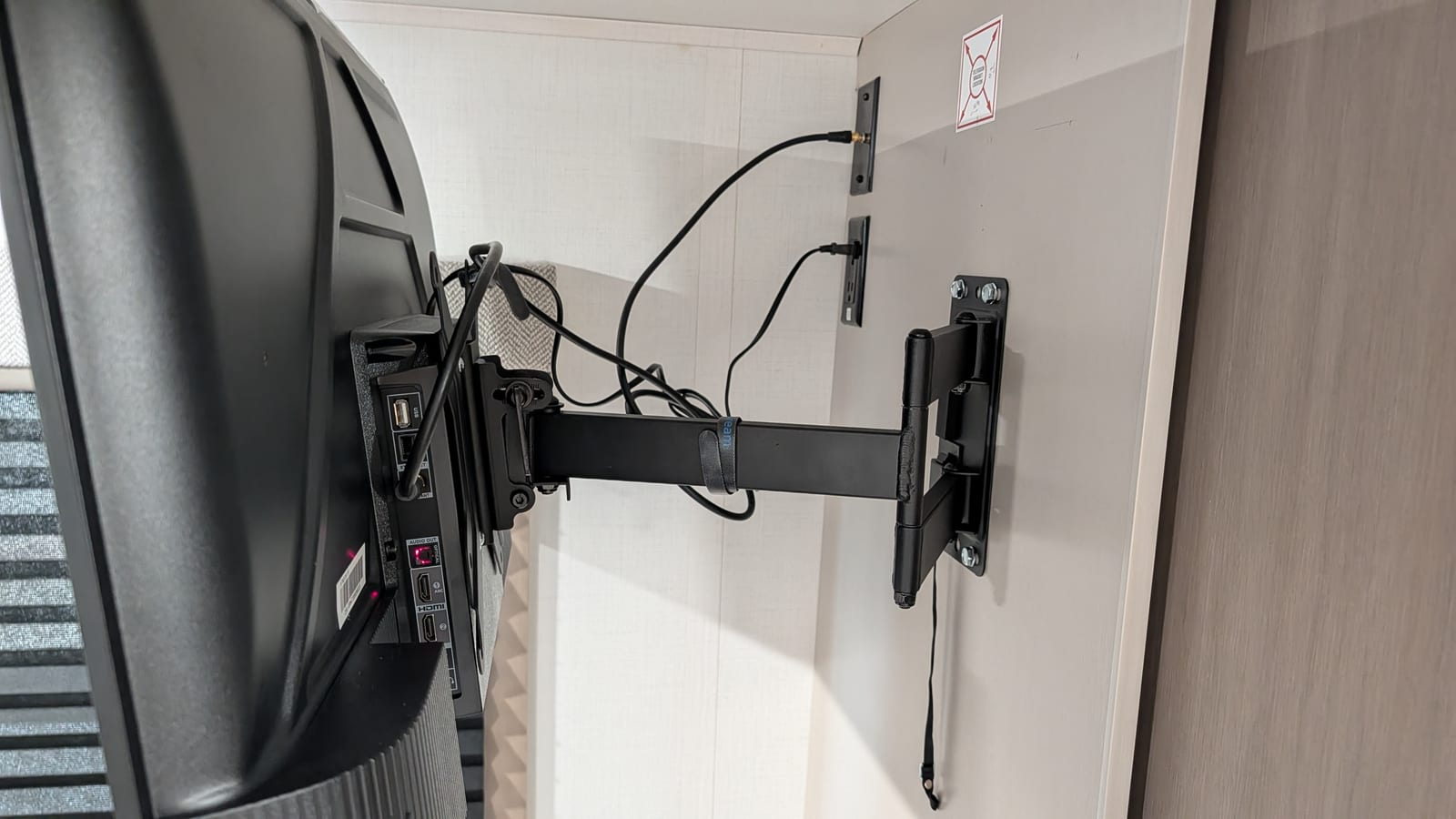 Side view of the TV pulled out from the wall on the double swing arm, showing the TV's rear ports with HDMI and coax, the cables running from the wall jacks, and a hook-and-loop strap around the arm.
