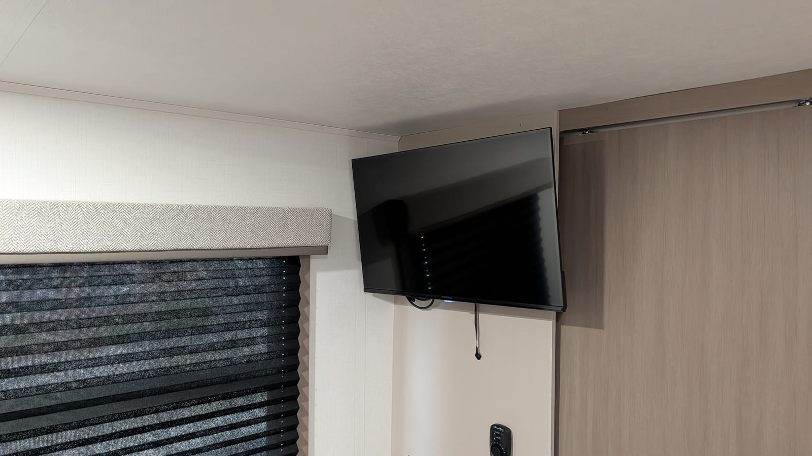A black 32-inch Insignia TV on a swing-arm mount, high in the corner of an RV bedroom, angled out from the wall beside a window with a fabric valance and pleated shade.