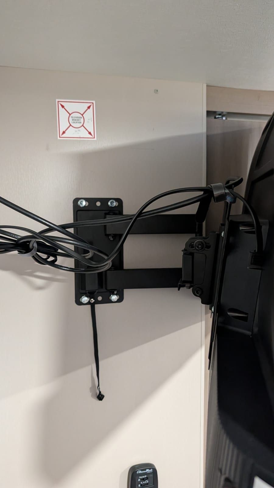 Portrait close-up of the black wall plate of the swing-arm mount, held by four bolts with washers, with a power cord and coax looped over the arm, a black lock-release strap hanging below the plate, and the back of the TV visible on the right.