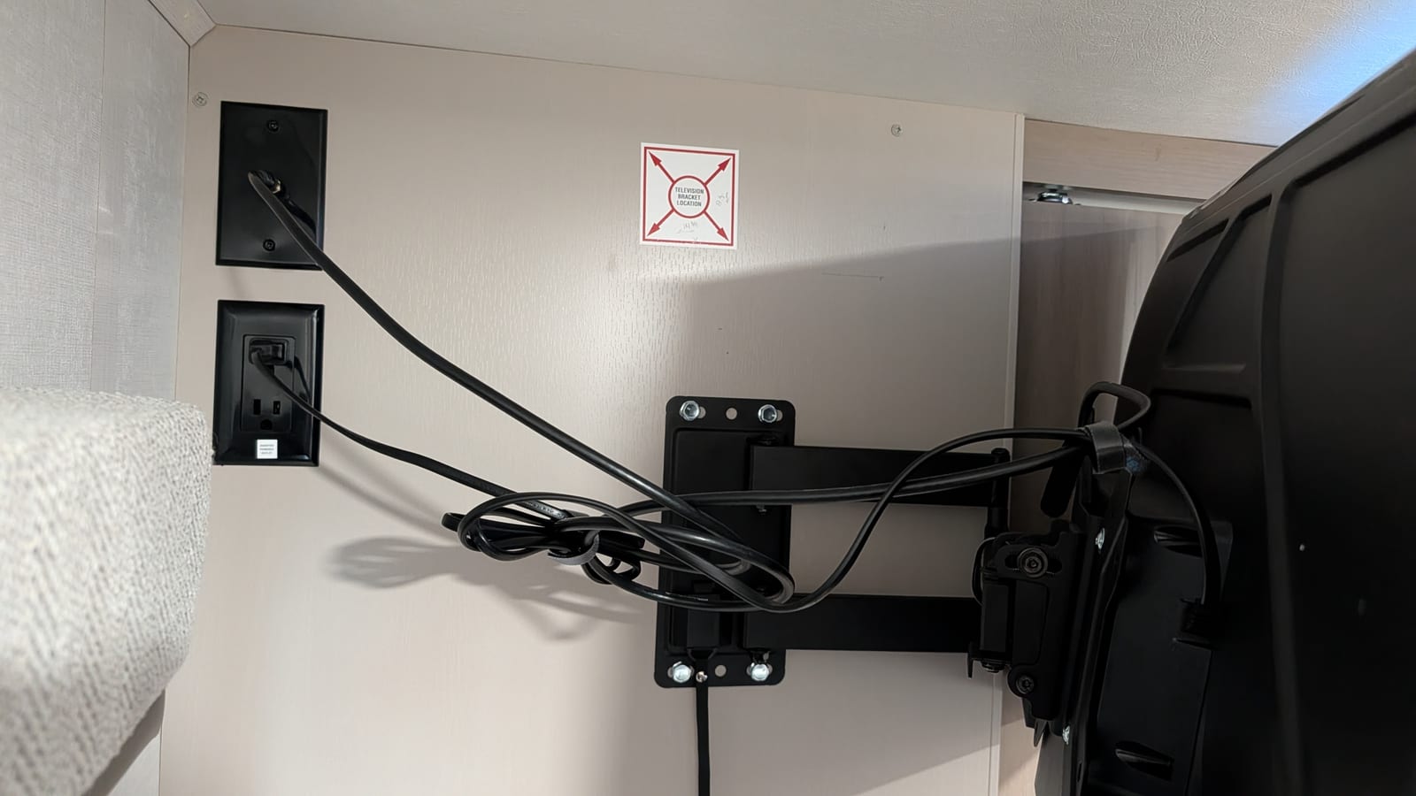 Close view of the RV bedroom wall showing a black coax wall plate and a black power outlet on the left, a red-and-white 'Television Bracket Location' sticker in the middle, and the black swing-arm mount's wall plate bolted below it with cables looped over the arm.
